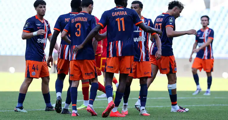 Soccer players in blue and orange jerseys form a huddle on the green field during a match. One player with number 11 stands central. Stadium seats are blurred in the background.