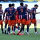 Soccer players in blue and orange jerseys form a huddle on the green field during a match. One player with number 11 stands central. Stadium seats are blurred in the background.