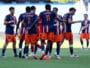 Soccer players in blue and orange jerseys form a huddle on the green field during a match. One player with number 11 stands central. Stadium seats are blurred in the background.