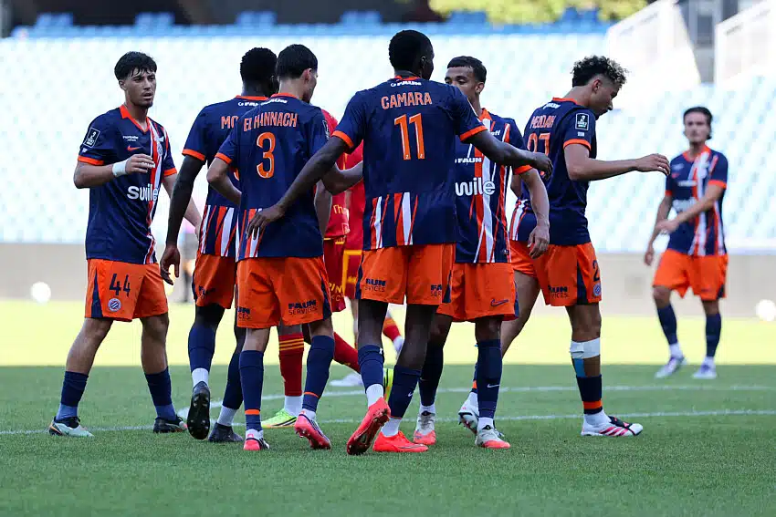 Joie | AllezPaillade.com Soccer players in blue and orange jerseys form a huddle on the green field during a match. One player with number 11 stands central. Stadium seats are blurred in the background.