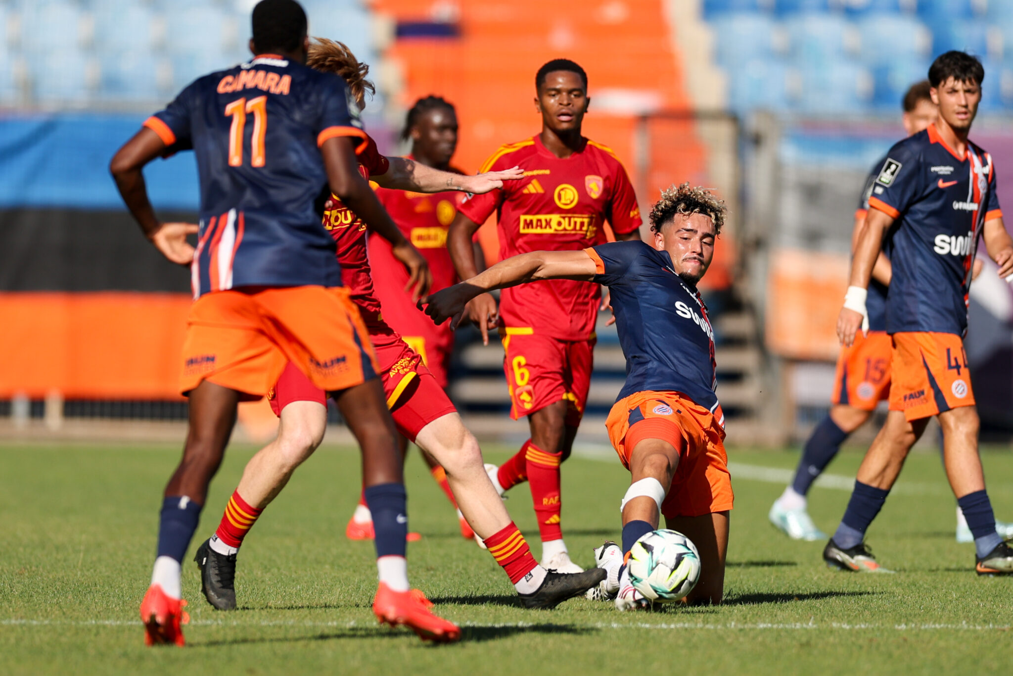 Soccer players in blue and red chase the ball on a grass field during a match, with stadium seating in the background.