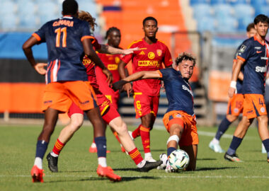 Soccer players in blue and red chase the ball on a grass field during a match, with stadium seating in the background.