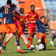 Soccer players in blue and red chase the ball on a grass field during a match, with stadium seating in the background.