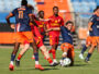 Soccer players in blue and red chase the ball on a grass field during a match, with stadium seating in the background.