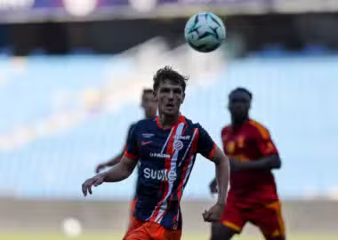 Soccer player in a blue-red striped kit looks up at a ball suspended above his head during a match, with teammates in the background.