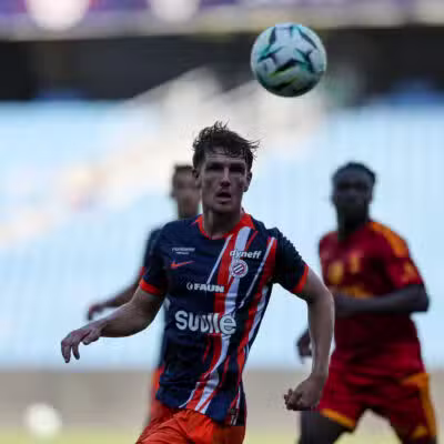 Soccer player in a blue-red striped kit looks up at a ball suspended above his head during a match, with teammates in the background.