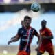 Soccer player in a blue-red striped kit looks up at a ball suspended above his head during a match, with teammates in the background.