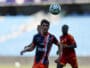 Soccer player in a blue-red striped kit looks up at a ball suspended above his head during a match, with teammates in the background.