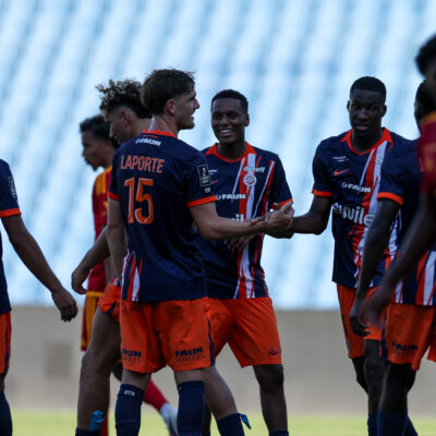 Group of young soccer players in blue and orange jerseys greet each other with handshakes and smiles on a grassy field.