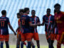 Group of young soccer players in blue and orange jerseys greet each other with handshakes and smiles on a grassy field.