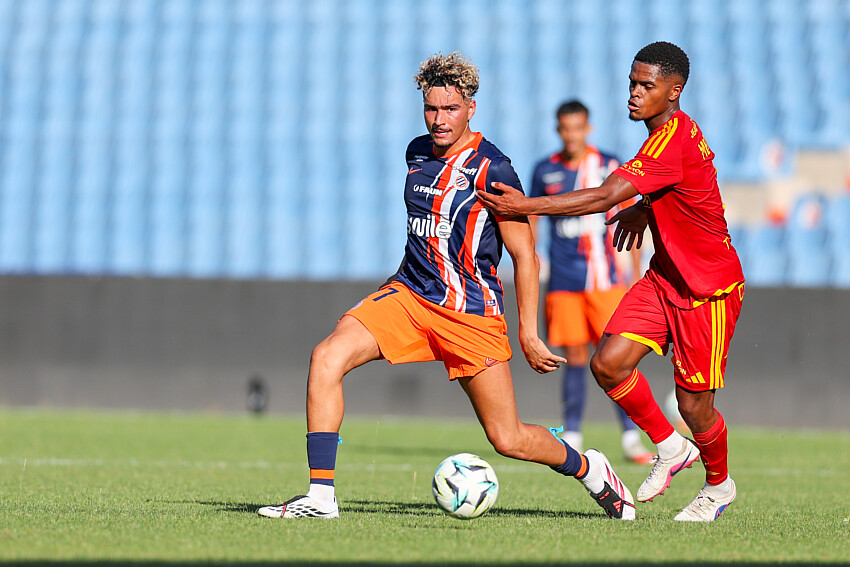 Montpellier Herault SC v Rodez Aveyron Football - Friendly Match 01082026 | AllezPaillade.com Two soccer players chase a ball on a grassy field, one wearing a blue and orange striped jersey and orange shorts, the other in red, as they contest possession.