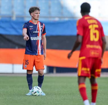 Soccer player in blue and orange kit with the ball at his feet, facing an opposing player in red in a stadium.