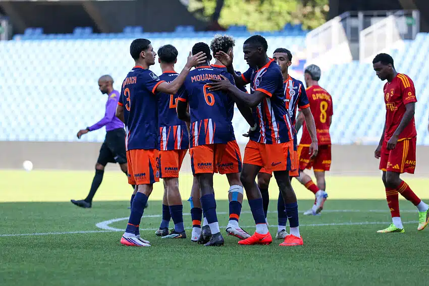 Joie | AllezPaillade.com Soccer players in blue and orange uniforms huddle in celebration on the field with teammates embracing each other amid a stadium backdrop.