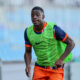 Young male runner in a neon green training vest and orange shorts sprinting on a track with a blurred stadium background.