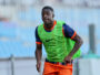 Young male runner in a neon green training vest and orange shorts sprinting on a track with a blurred stadium background.