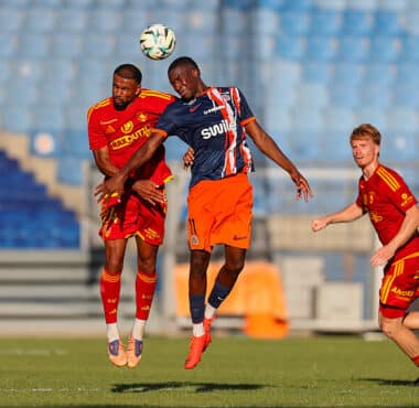 Three soccer players jump to head the ball during a match on a green field, with blue stadium seats in the background.