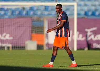 Soccer player in a blue and orange striped jersey walking on a grass field with a goal in the background in sunny stadium light.