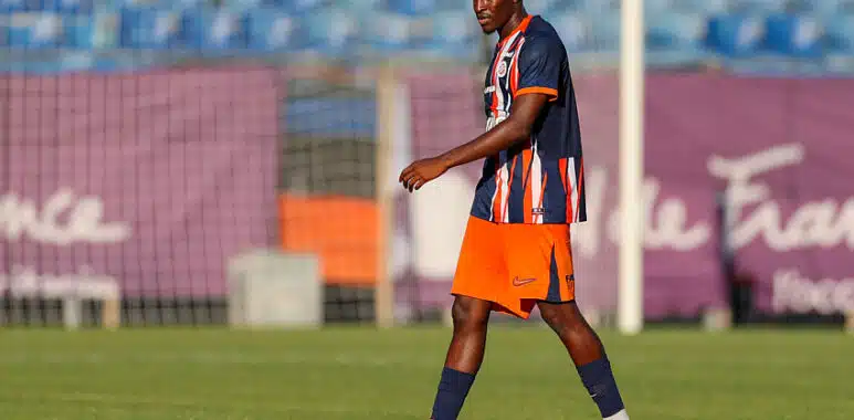 Camara | AllezPaillade.com Soccer player in a blue and orange striped jersey walking on a grass field with a goal in the background in sunny stadium light.