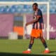 Soccer player in a blue and orange striped jersey walking on a grass field with a goal in the background in sunny stadium light.