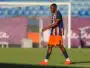 Soccer player in a blue and orange striped jersey walking on a grass field with a goal in the background in sunny stadium light.