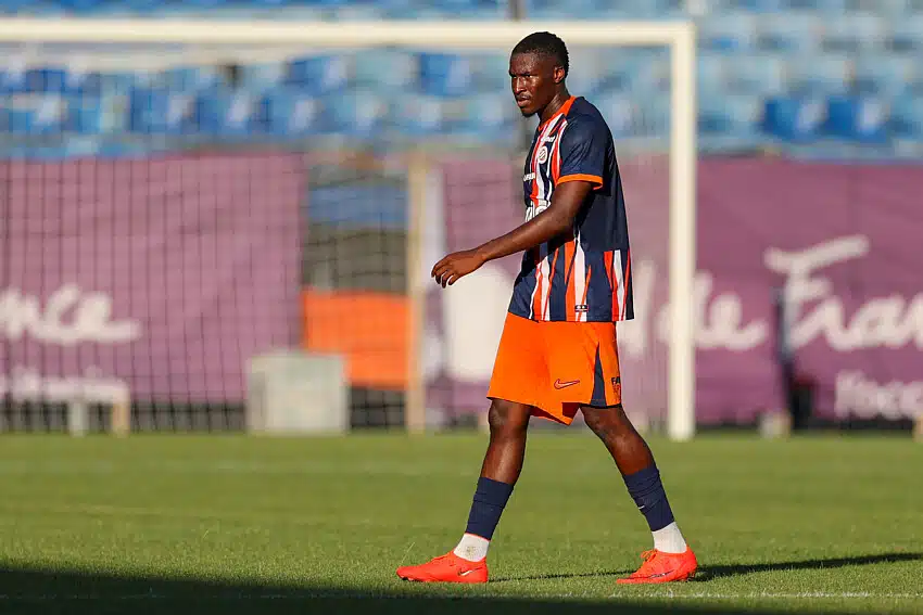 Camara | AllezPaillade.com Soccer player in a blue and orange striped jersey walking on a grass field with a goal in the background in sunny stadium light.