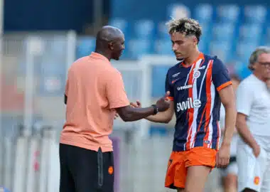 Soccer player in a blue and orange striped kit shakes hands with a man in a peach shirt on the sideline, with stadium seating in the background.