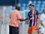 Soccer player in a blue and orange striped kit shakes hands with a man in a peach shirt on the sideline, with stadium seating in the background.
