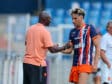 Soccer coach and player shake hands on the field after a training session; man in peach polo and another in blue-orange striped kit. Keep it concise and clear for screen readers, with setting on the pitch and blurred stands in the background.