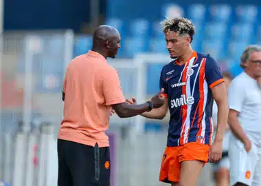 Soccer coach and player shake hands on the field after a training session; man in peach polo and another in blue-orange striped kit. Keep it concise and clear for screen readers, with setting on the pitch and blurred stands in the background.