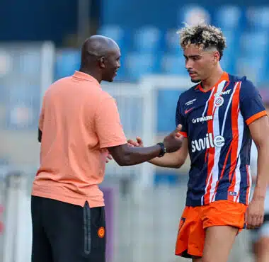 Soccer coach and player shake hands on the field after a training session; man in peach polo and another in blue-orange striped kit. Keep it concise and clear for screen readers, with setting on the pitch and blurred stands in the background.