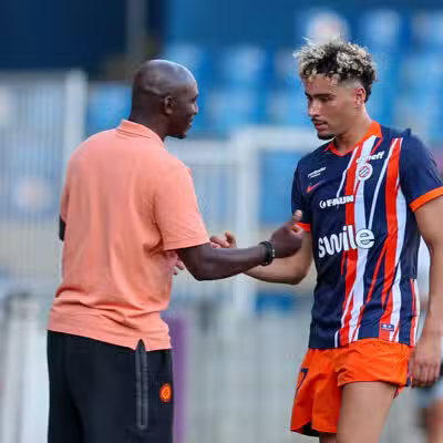 Soccer coach and player shake hands on the field after a training session; man in peach polo and another in blue-orange striped kit. Keep it concise and clear for screen readers, with setting on the pitch and blurred stands in the background.