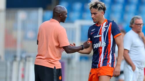 Soccer coach and player shake hands on the field after a training session; man in peach polo and another in blue-orange striped kit. Keep it concise and clear for screen readers, with setting on the pitch and blurred stands in the background.