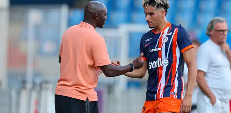 Meddah | AllezPaillade.com Soccer coach and player shake hands on the field after a training session; man in peach polo and another in blue-orange striped kit. Keep it concise and clear for screen readers, with setting on the pitch and blurred stands in the background.