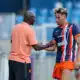Soccer coach and player shake hands on the field after a training session; man in peach polo and another in blue-orange striped kit. Keep it concise and clear for screen readers, with setting on the pitch and blurred stands in the background.