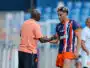 Soccer coach and player shake hands on the field after a training session; man in peach polo and another in blue-orange striped kit. Keep it concise and clear for screen readers, with setting on the pitch and blurred stands in the background.