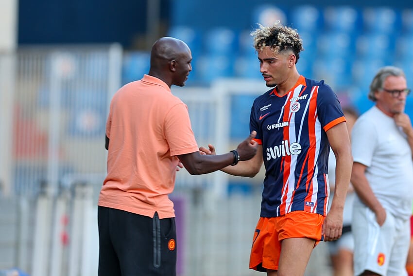 Soccer coach and player shake hands on the field after a training session; man in peach polo and another in blue-orange striped kit. Keep it concise and clear for screen readers, with setting on the pitch and blurred stands in the background.