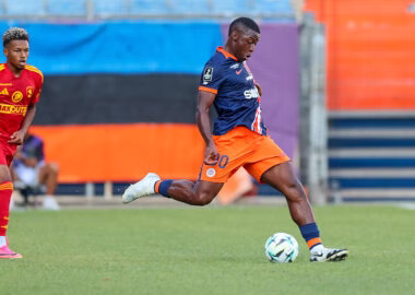 Ayem | AllezPaillade.com Soccer player in navy and orange kit strikes the ball on a grassy field as a rival in red/yellow watches nearby.