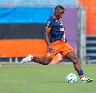 Soccer player in navy and orange kit strikes the ball on a grassy field as a rival in red/yellow watches nearby.