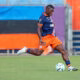 Soccer player in navy and orange kit strikes the ball on a grassy field as a rival in red/yellow watches nearby.