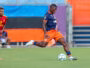 Soccer player in navy and orange kit strikes the ball on a grassy field as a rival in red/yellow watches nearby.