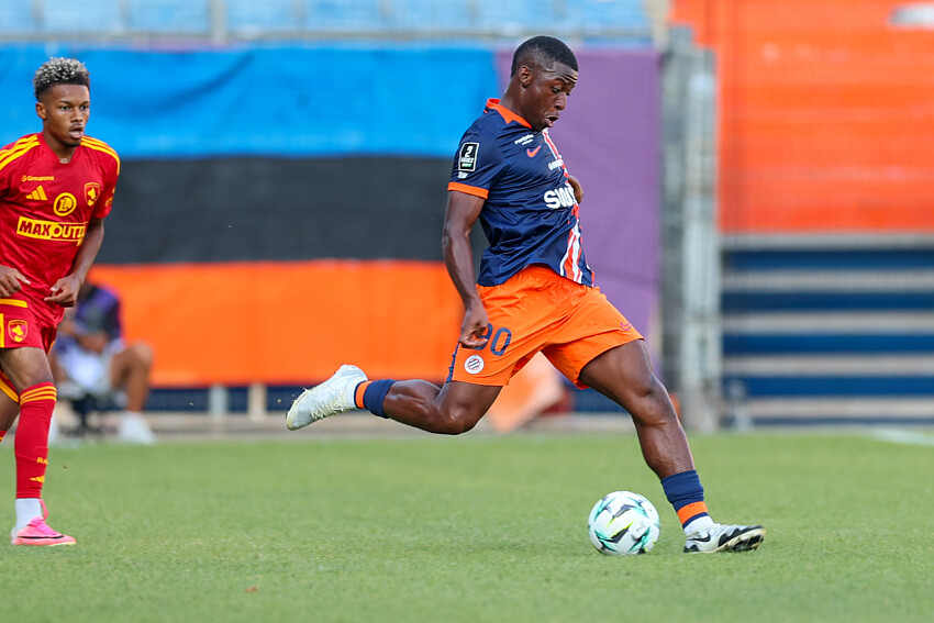 Ayem | AllezPaillade.com Soccer player in navy and orange kit strikes the ball on a grassy field as a rival in red/yellow watches nearby.
