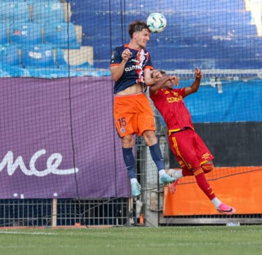 Two soccer players jump for a header near the goal, one in navy/orange and the other in red, with a net and blue seats behind them.