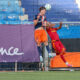 Two soccer players jump for a header near the goal, one in navy/orange and the other in red, with a net and blue seats behind them.