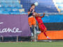Two soccer players jump for a header near the goal, one in navy/orange and the other in red, with a net and blue seats behind them.