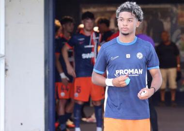 Young male soccer player in blue jersey stepping out of the tunnel, teammates in orange uniforms behind him.