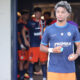 Young male soccer player in blue jersey stepping out of the tunnel, teammates in orange uniforms behind him.