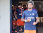 Young male soccer player in blue jersey stepping out of the tunnel, teammates in orange uniforms behind him.