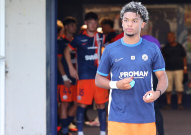 Young male soccer player in a blue jersey holding a ball, standing near a tunnel with teammates in orange shorts in the background.