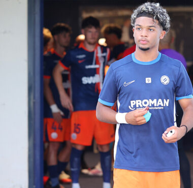 Young male soccer player in a blue jersey holding a ball, standing near a tunnel with teammates in orange shorts in the background.