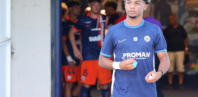 Mégnan | AllezPaillade.com Young male soccer player in a blue jersey holding a ball, standing near a tunnel with teammates in orange shorts in the background.